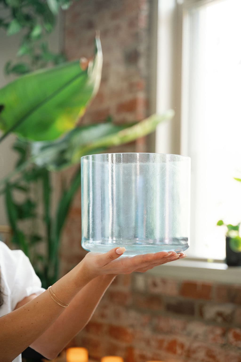 Hands holding a clear crystal singing bowl up to soft window light beside a leafy plant